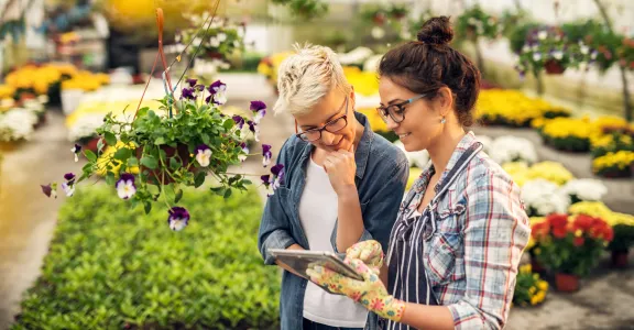 Deux femmes dans une jardinerie, absorbées dans une tablette, discutant de plantes et de conseils de jardinage.